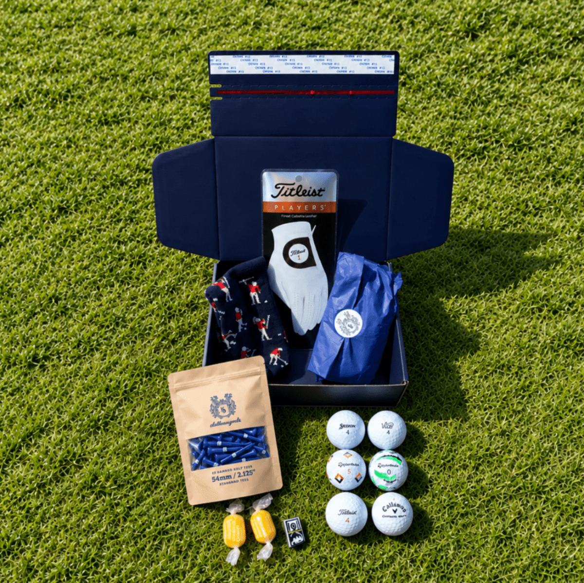 Members Mystery Box on grass: navy Clubhouse Goods box with a Titleist glove, golf balls, tees and sweets