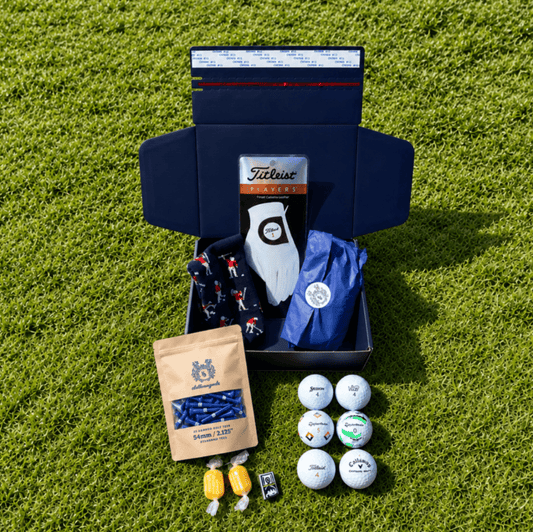 Members Mystery Box on grass: navy Clubhouse Goods box with a Titleist glove, golf balls, tees and sweets